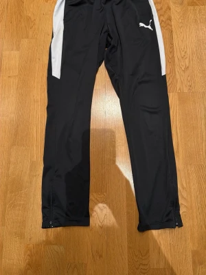 Svarta träningsbyxor från Puma - Snygga svarta träningsbyxor från Puma med vita detaljer längs sidorna och dragkedjor vid bensluten. Byxorna har en sportig passform och är tillverkade i ett lätt och funktionellt material som passar perfekt till träning eller uppvärmning.