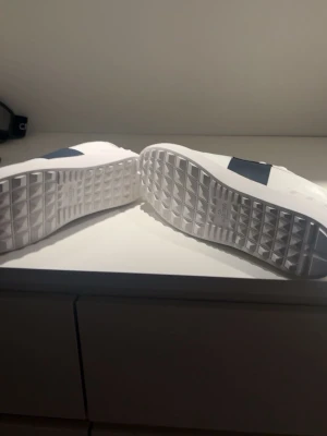 Vita sneakers från Valentino Garavani - Snygga vita sneakers från Valentino Garavani med bred svart detalj över mitten och guldfärgad logga på insidan. Skorna har vita snören, robust sula med rutmönster undertill och klassisk låg profil. Perfekta för dig som gillar exklusiv och stilren design. Ej använda och de kommer till en box med 