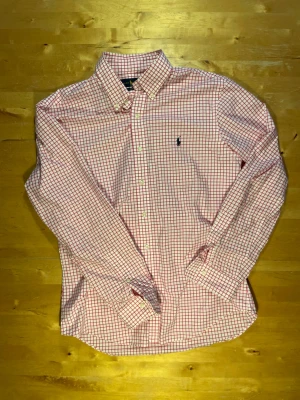 Rutig skjorta från Ralph Lauren - Klassisk rutig skjorta från Ralph Lauren i vitt och rött med blå logga på bröstet. Skjortan har lång ärm, knappar framtill och button-down krage. Tillverkad i mjuk bomull, perfekt för en clean och stilren look.