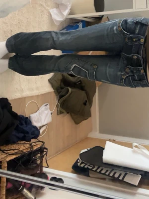 Lågmidjade jeans - Säljer ett par blåa jeans med coola slitningar och en unik, detaljerad ficka på låret. Jeansen har låg midja, raka ben och kontrastsömmar. Perfekta för dig som vill ha en avslappnad men ändå trendig look.