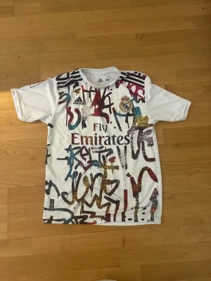 Real Madrid tröja Vini Jr. graffiti - Säljer en vit Real Madrid retro tröja från Adidas med coolt graffiti-mönster i flera färger och klubbmärke på bröstet, den passar för både 176 och S. På ryggen står det VINI JR. och nummer 20. Perfekt för dig som älskar fotboll och streetstyle. Pris kan diskuteras!