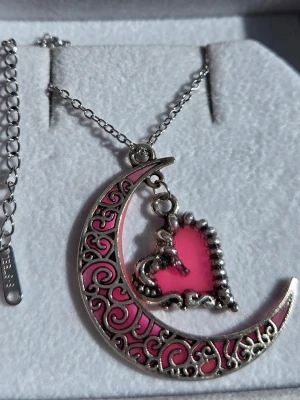 Halsband med måne och hjärta - Självlysande  - Unikt halsband i silverfärgad rostfritt stål med en halvmåne dekorerad med rosa och lila detaljer som lyser i mörkret. I mitten hänger ett rosa hjärta med dekorativ kant. Vanligt dagsljus och solljus aktiverar effekten i hänget. Perfekt för dig som gillar magiska och romantiska smycken. Både kedjan och hänget tål vatten och missfärgas inte. Hängets storlek är ca 3 cm och kedjan 45 cm + 5 cm förlängningskedja.