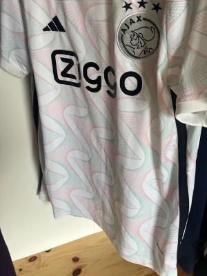 Ajax Amsterdam matchtröja vit - Säljer en Ajax Amsterdam matchtröja från Adidas i vitt med pastellfärgade mönster och svarta detaljer. Tröjan har klubbmärke, sponsorlogga och tre stjärnor på bröstet. Tillverkad i lätt och ventilerande material, perfekt för fotboll eller träning.