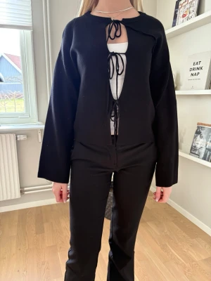 Svart cardigan med knytband - Trendig svart cardigan med vida ärmar och tre knytband framtill. Öppen modell som ger en avslappnad look, perfekt att styla över ett linne eller t-shirt. Mjuk och skön kvalitet som passar till många outfits.
