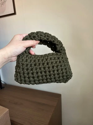 Olivgrön virkad handväska - Unik handväska i olivgrönt, grovt virkat garn. Väskan har en kompakt, rektangulär form med ett matchande handtag i samma material. Perfekt för dig som gillar handgjorda accessoarer med personlig touch. Ingen synlig logga eller märke.