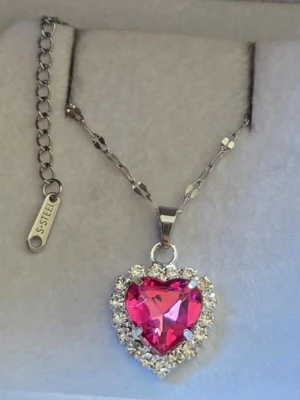 Halsband med hjärta och strass - Snyggt halsband i rostfritt stål med ett stort hjärtformat rosa hänge omgivet av gnistrande klara stenar. Kedjan har en unik länkdesign och justerbar längd. Perfekt för dig som gillar statement-smycken och vill sticka ut.
