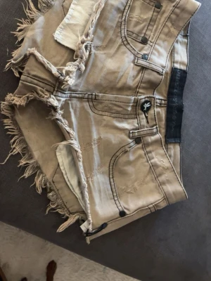 Low waist One Teaspoon shorts - Säljer ett par beige/gröna  shorts från One Teaspoon i storlek w24 med råa, fransiga kanter och slitna detaljer. De har klassiska fickor fram och bak, knappstängning och en cool logotyp på linningen. Perfekta för en avslappnad och trendig look.