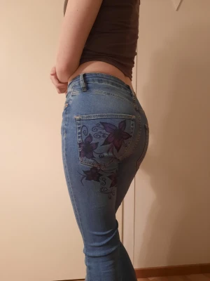 Blå bootcut jeans med blommor - Snygga ljusblå bootcut jeans med lila och svarta blommor målade på bakfickan och höften. Jeansen har klassisk femficksdesign och är tillverkade i stretchigt denimtyg för en bekväm passform. Perfekta för dig som vill sticka ut med en unik detalj.