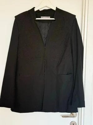 Svart stilig kavaj från Gina Tricot - Stilren svart kavaj med axelvaddar från Gina Tricot med diskret mönstrad struktur upptill. Rak passform, öppen framtill utan knappar och två fickor. Perfekt för en clean och modern look.