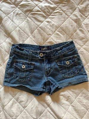 Blå lågmidjande jeansshorts - Snygga blå lågmidjande vintage jeansshorts från Angels med fina fickor med knappar både fram och bak. Har ingen utskriven storlek men dom är 34cm i midjan platta, jag är 84cm runt i höften där shortsen sitter. Dom passar bra på mig då. 