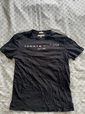 Svart Tommy Hilfiger t-shirt - Snygg svart t-shirt från Tommy Hilfiger med klassisk logga och texten 'EST. 1985' på bröstet. T-shirten har rund halsringning och korta ärmar. Tillverkad i mjuk bomull som känns skön mot huden. Perfekt till jeans eller shorts.