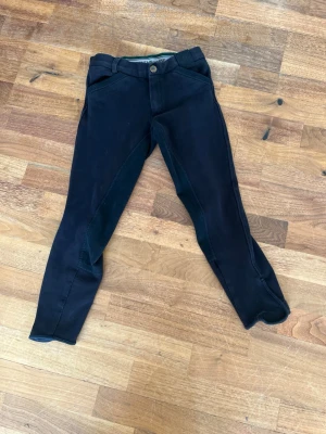 Svarta ridbyxor från Mountain Horse - Säljer ett par svarta ridbyxor från Mountain Horse med helskoning i mockaimitation. Byxorna har bälteshällor, knapp och dragkedja framtill samt förstärkta partier på insidan av benen och bak. 