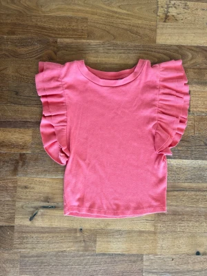 Rosa topp med volangärmar - Supersöt rosa topp med ribbad struktur och stora volangdetaljer längs ärmarna. Perfekt för dig som vill sticka ut lite extra! Toppen har rund halsringning och är tillverkad i mjuk bomull för skön känsla hela dagen.