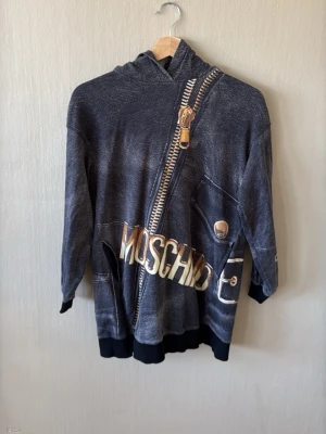 Moschino hoodie med dragkedjeprint - Cool hoodie från Moschino Teen i mörkgrått med ett stort, detaljerat dragkedjeprint och Moschino-logga över bröstet. Hoodien har huva och svarta muddar vid ärmslut och nederkant. Materialet känns mjukt och bekvämt, perfekt för en avslappnad streetwear-look.