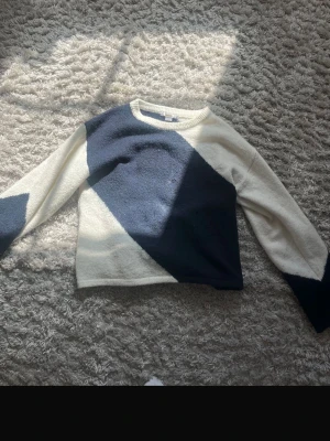 Stickad tröja med blåvitt mönster - Säljer en stickad tröja i vitt med stora mörkblå diagonala block på både fram- och baksida samt ärmar. Tröjan har rund halsringning och långa ärmar. 
