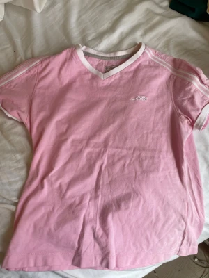 Rosa t-shirt från Lotto med v-ringning - Säljer en rosa t-shirt från Lotto med vit v-ringning och vita detaljer vid ärmslut. T-shirten har korta ärmar och är tillverkad i mjuk bomull. Diskret Lotto-logga broderad på bröstet. Perfekt för dig som gillar sportiga och stilrena plagg.