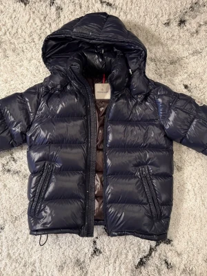 Moncler Maya - Snygg mörkblå dunjacka från Moncler med glansig finish, huva och två dragkedjeförsedda fickor. Jackan har ett klassiskt Moncler-märke på ärmen och quiltad design. Perfekt för kalla vinterdagar och riktigt trendig look. Allt från köpet i Moncler butiken medföljer. 