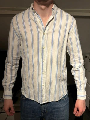 Randig skjorta i bomull med blå toner - Säljer en vit skjorta med blå och beige ränder. Skjortan har klassisk krage, knappar framtill och långa ärmar. Tillverkad i mjuk bomull och har en avslappnad passform som passar perfekt till jeans. Stor i storleken 