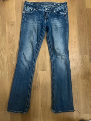 Blå bootcut jeans från Miss Me - Säljer ett par blå bootcut jeans från Miss Me med snygga slitningar och kontrastsömmar. Jeansen har fem fickor, silverfärgade knappar och dragkedja. Perfekta för dig som gillar en avslappnad men trendig look.