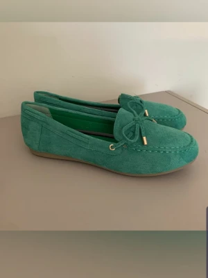 Gröna mockaloafers med rosett nya - Snygga loafers i grön mocka med dekorativ rosett framtill och guldfärgade detaljer. Skorna har en klassisk seglarsko-look med låg sula i gummi och stilren design. Perfekta för dig som vill ha en chill men ändå trendig stil.