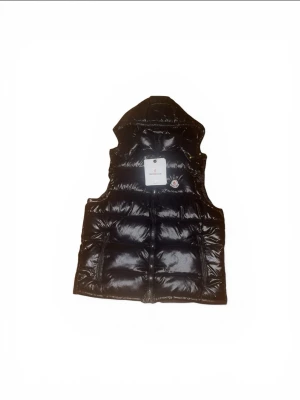 Svart dunväst från Moncler - Snygg svart dunväst från Moncler med glansig finish och klassisk logotyp på bröstet. Västen har huva och dragkedja framtill samt två fickor. Perfekt för lager på lager och streetwear-stil.