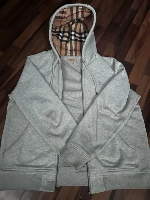 Grå hoodie från Burberry med mönstrad huva - Snygg grå zip-hoodie från Burberry med den klassiska rutiga mönstringen i beige, svart, rött och vitt på insidan av huvan. Hoodien har dragsko i huvan, fickor framtill och är tillverkad i mjukt bomullsmaterial. Perfekt för en chill och stilren look. Tagen är Xl men sitter mer som L