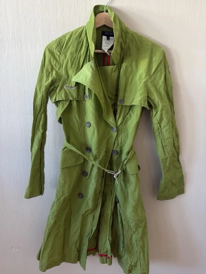 Grön trenchcoat från Exté, strl It 44 - Snygg grön trenchcoat från Exté med dubbelknäppning, bred krage och bälte i midjan. Tillverkad i Italien av polyamid och metallfiber för en unik look. Fickor med lock och coola detaljer. Perfekt för dig som vill sticka ut med din stil. Längd: 70 cm 