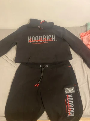 Svart Hoodrich loungewear set, den som köper direkt får 25% Rabbat  - Säljer ett svart loungewear set från Hoodrich med matchande hoodie och byxor. Setet har tryckt logga i vitt och rött på både tröjan och byxorna, samt röda detaljer på snören och dragkedjor. Perfekt för chill dagar hemma eller streetwear vibes. Tröjan har storlek S och byxorna M