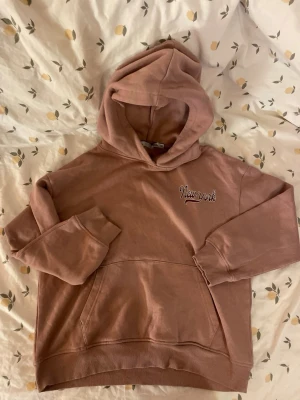 Rosa hoodie med New York-tryck Barn 140 - Säljer en mjuk rosa hoodie från Lager 157 med huva och känguruficka. På bröstet står det 'New York' i vit och brun text. Perfekt för chill dagar och enkel att matcha med jeans eller leggings. Tillverkad i bomullsmix. Barn storlek 140