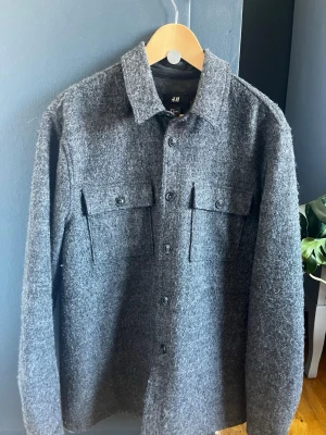 Grå overshirt från H&M - Stilren grå overshirt från H&M i mjukt, ull-liknande material. Klassisk skjortkrage, två bröstfickor med lock och knappar framtill. Perfekt lager-på-lager-plagg med avslappnad passform och långa ärmar.
