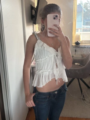 Vit omlott linnetopp med volanger - Säljer en vit linnetopp med omlott-design och volanger. Toppen har smala axelband, knytning framtill och är croppad med plisserade detaljer. Perfekt för sommarens alla tillfällen, superfräsch och trendig.