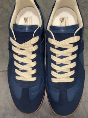 Maison Margiela - Blå sneakers från Maison Margiela i storlek 41, med beige snören och detaljer i mocka och läder. Klassisk låg modell med vit innersula och slitstark gummisula som passar till det mesta. Skorna är använda men däremot i bra skick, med gott om liv kvar. Då menar jag att skorna på bilden är när dem var nya och nu har dem används och ser inte lika nya ut.