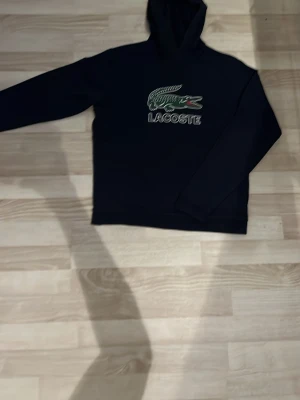 Mörkblå hoodie från Lacoste - Snygg mörkblå hoodie från Lacoste med klassisk krokodillogga och text på bröstet. Hoodien har huva och långa ärmar, perfekt för en avslappnad och sportig stil.