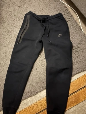 Svarta Nike mjukisbyxor med dragkedja - Svarta mjukisbyxor från Nike med smal passform, dragsko i midjan och en stor dragkedjeficka på ena benet. Byxorna har Nike-logga på låret och är tillverkade i mjukt material för maximal komfort. Nike tech storlek M ej använt många gånger för stora för mig 