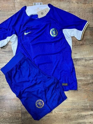 Chelsea FC matchställ Nike blå - Chelsea FC matchställ i blått med vita detaljer. Tröjan har klubbmärke och Nike-logga på bröstet, samt guldkant på ärmen. Shortsen är blå med Chelsea-logga. Perfekt för fotbollsträning eller match. Materialet är lätt och andas.