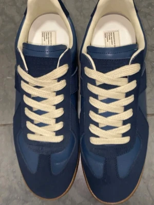 Maison Margiela - Blå sneakers från Maison Margiela i storlek 41, med beige snören och detaljer i mocka och läder. Klassisk låg modell med vit innersula och slitstark gummisula som passar till det mesta. Skorna är använda men däremot i bra skick, med gott om liv kvar. Då menar jag att skorna på bilden är när dem var nya och nu har dem används och ser inte lika nya ut.