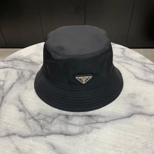 Svart bucket hat - Stilren svart bucket hatmed klassisk form och ikonisk metallogo framtill. Hatten är tillverkad i ett glansigt syntetmaterial som ger en modern känsla och passar perfekt till streetwear eller en trendig look.