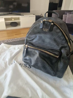 Guess Ryggsäck - Cool svart ryggsäck från Guess med subtilt mönster och guldfärgade dragkedjor. Ryggsäcken har ett stort huvudfack, en mindre ficka framtill och justerbara axelband. Materialet är konstläder och logotypen är i metall på framsidan.