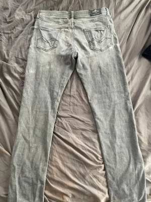 Grå Replay jeans - Snygga grå slim fit jeans från Replay med klassisk femficksdesign och ljus tvätt. Jeansen har raka ben och detaljerade bakfickor med sömmar. Perfekta för en avslappnad och trendig stil. Passar w30-w31.