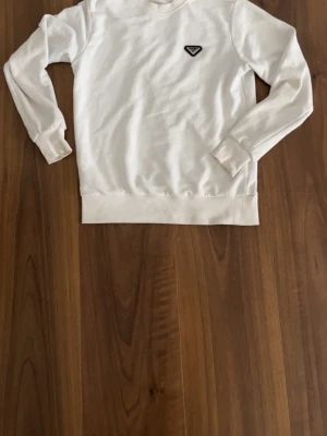 Vit sweatshirt från Prada - Vit sweatshirt från Prada med klassisk rund hals och långa ärmar. Tröjan har ett snyggt metalliskt Prada-logoemblem på bröstet och ribbade muddar vid ärmslut och nederkant. Perfekt för en clean och stilren look.