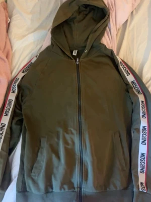 Olivgrön hoodie från Moschino - Snygg olivgrön hoodie från Moschino med dragkedja och huva. På ärmarna finns vita band med Moschino-logga i svart och röd kant. Mjukt material och två fickor framtill. Perfekt för en trendig och avslappnad stil.