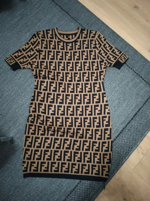 Beige och svart Fendi klänning - Cool kortärmad klänning från Fendi med ikoniskt FF-mönster i beige och svart. Klänningen har rund halsringning och är figurnära med ribbade kanter vid ärmslut och nederkant. Perfekt för dig som vill sticka ut med designerstil.