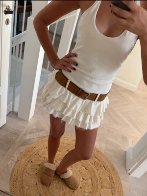 Vit volangkjol med bälte - Supersöt vit kortkjol med flera lager volanger och ett snyggt brunt bälte i midjan. Kjolen har en feminin och lekfull stil, perfekt för dig som gillar trendiga och unika plagg. Passar perfekt till sommarens alla outfits.