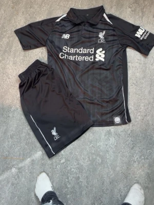 Liverpool svart träningsset New Balance - Säljer ett svart träningsset från Liverpool FC med kortärmad tröja och shorts. Tröjan har vit logga, sponsortryck och krage, medan shortsen har elastisk midja och vit Liverpool-logga. Materialet är lätt och andas, perfekt för fotboll eller träning.