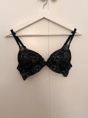 Svart spets-bh med silvriga detaljer - Snygg svart bh med vadderade kupor (ej push-up). Bh:n har ett elegant spetsmönster i svart och silver, samt hakknäppning bak. Justerbara axelband för bästa passform. Perfekt för dig som gillar en stilren och feminin look.