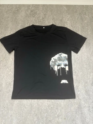 Svart t-shirt med unik tryck - Cool svart t-shirt med ett stort, konstnärligt tryck på framsidan som föreställer en droppande, silvrig design. T-shirten har rund hals och korta ärmar, perfekt för dig som vill sticka ut med en edgy look.