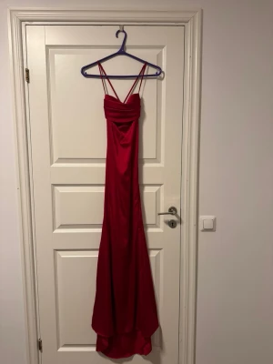 Röd lång klänning med smala axelband - Säljer en elegant röd långklänning med smala axelband och draperad överdel. Klänningen har en figurnära passform och är tillverkad i ett glansigt material som ger en lyxig känsla. Perfekt för bal eller festliga tillfällen.