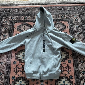 Grå hoodie från Stone Island - Snygg grå hoodie från Stone Island med dragkedja framtill och klassisk logopatch på vänster ärm. Hoodien har två fickor fram, ribbade muddar och justerbar huva med snören. Perfekt för en avslappnad och trendig stil.