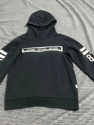 Svart hoodie med vita detaljer - Snygg svart hoodie från WOKXI med vita tryckdetaljer på bröstet och ärmarna. Huva och ribbade muddar vid ärmslut och nederkant. Perfekt för en avslappnad streetwear-look.