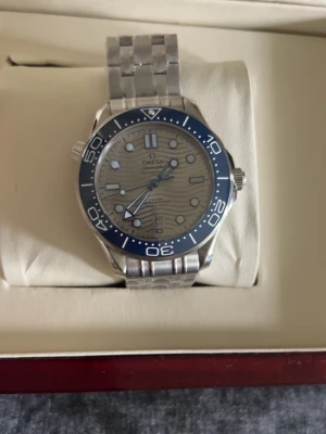 Omega Seamaster silverklocka - Säljer en Omega Seamaster herrklocka i silver med blå vridring och länkarmband i metall. Urtavlan är ljus med vågmönster och tydliga indexmarkeringar. Klockan levereras i originalbox med tillhörande certifikat och dokumentation.